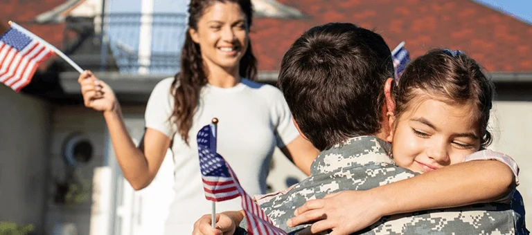 A soldier hugs a child in front of a home, while another person waves miniature American flags in a joyful reunion.