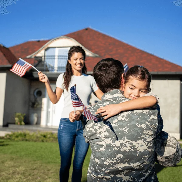 A soldier hugs a child in front of a home, while another person waves miniature American flags in a joyful reunion.