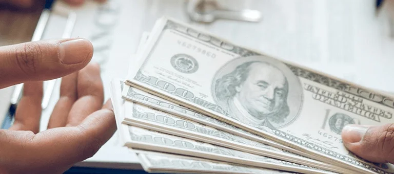 Hands exchanging cash over a document, with a small model house and key in the background, signifying a real estate transaction.