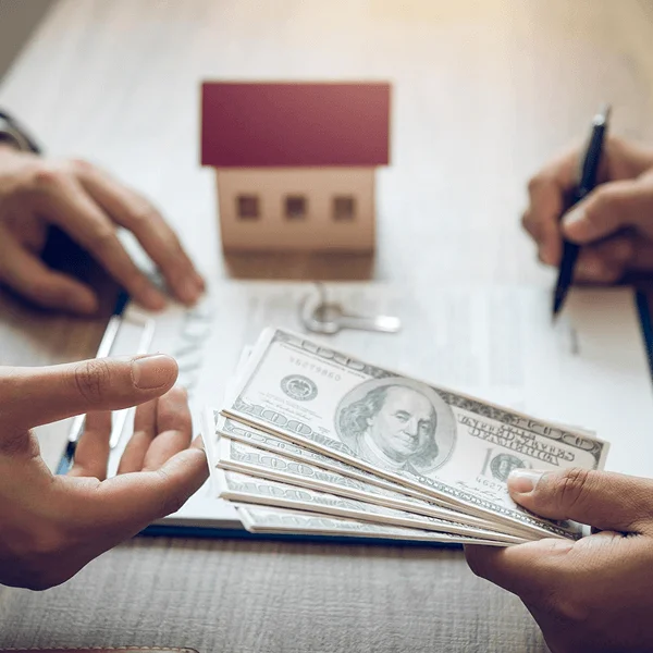 Hands exchanging cash over a document, with a small model house and key in the background, signifying a real estate transaction.