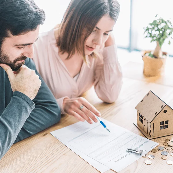 A couple sits at a wooden table reviewing paperwork, with a small model house and coins nearby, indicating a discussion about homeownership.