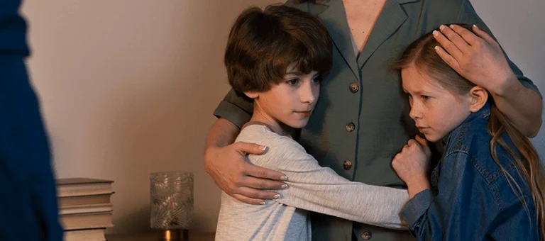 A woman stands with two children, embracing them closely, while a figure in the foreground appears to be watching them. Books are stacked nearby.