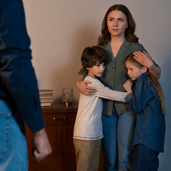 A woman stands with two children, embracing them closely, while a figure in the foreground appears to be watching them. Books are stacked nearby.