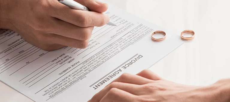 A hand signs a divorce agreement on a table with two wedding rings beside it, symbolizing the end of a marriage.
