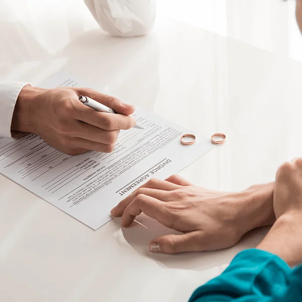 A hand signs a divorce agreement on a table with two wedding rings beside it, symbolizing the end of a marriage.