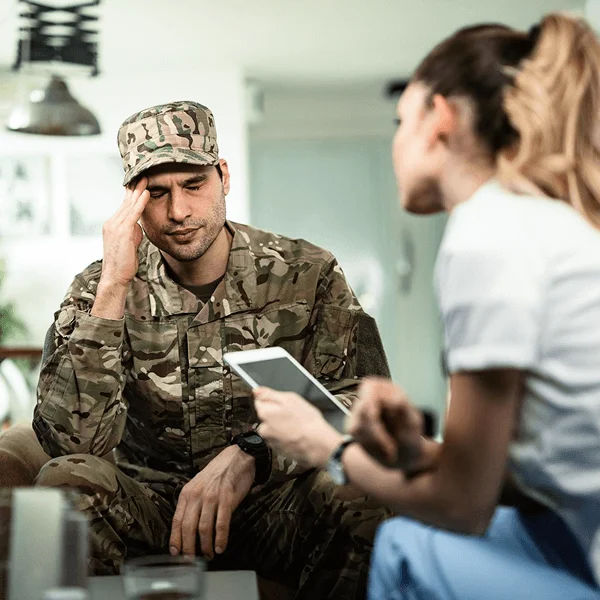 A soldier in military attire sits with a pained expression, while a healthcare professional engages with him, holding a tablet.