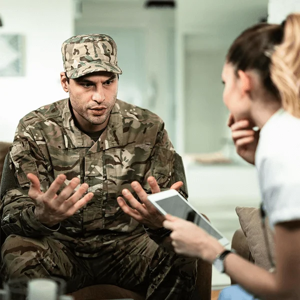 A soldier in military fatigues sits on a couch, animatedly speaking to a woman holding a tablet, engaged in a serious conversation.