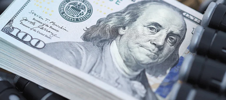 A stack of hundred-dollar bills lies on a camouflage surface, surrounded by several black-tipped projectiles, suggesting a tense atmosphere.
