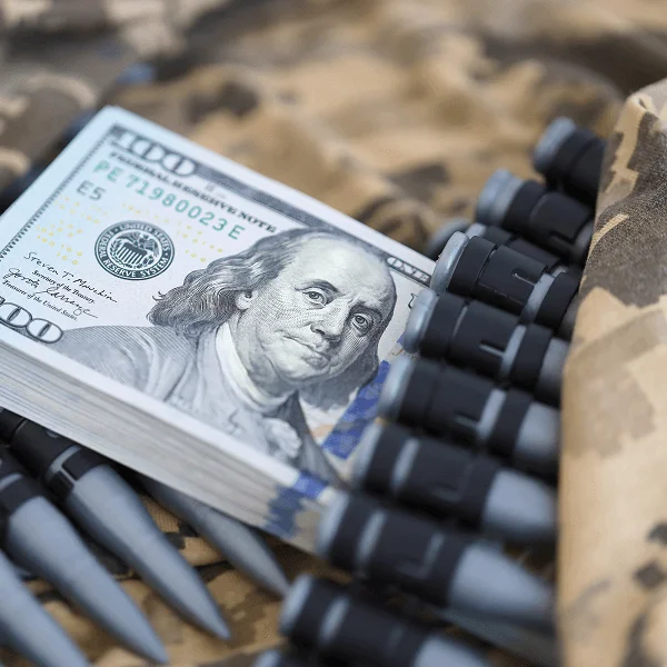 A stack of hundred-dollar bills lies on a camouflage surface, surrounded by several black-tipped projectiles, suggesting a tense atmosphere.