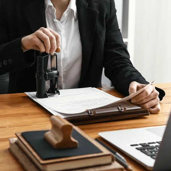 A businessman in a suit using a seal stamp on a document while holding another paper, seated at a wooden desk.