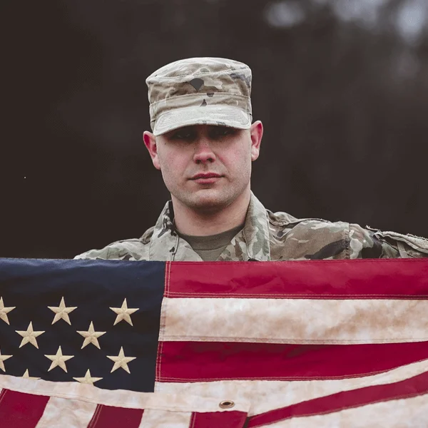 A soldier in military uniform proudly holds an American flag with stars and stripes against a dark backdrop.