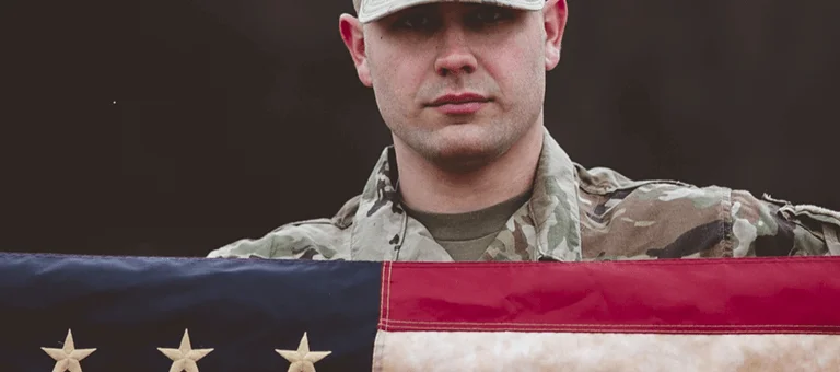 A soldier in military uniform proudly holds an American flag with stars and stripes against a dark backdrop.