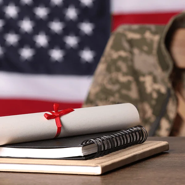 A rolled diploma tied with a red ribbon sits atop a notebook, with a soldier in a camouflaged uniform and American flag in the background.