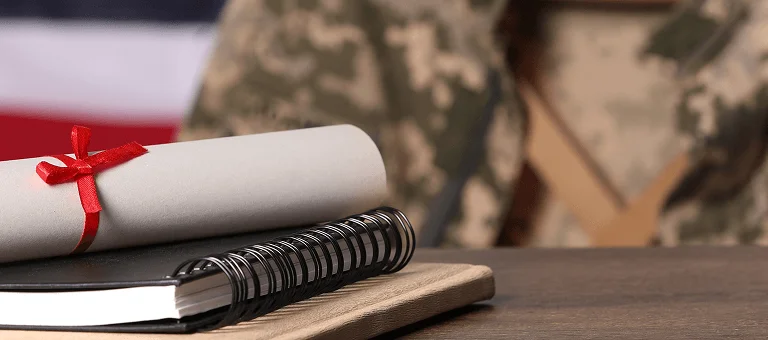 A rolled diploma tied with a red ribbon sits atop a notebook, with a soldier in a camouflaged uniform and American flag in the background.