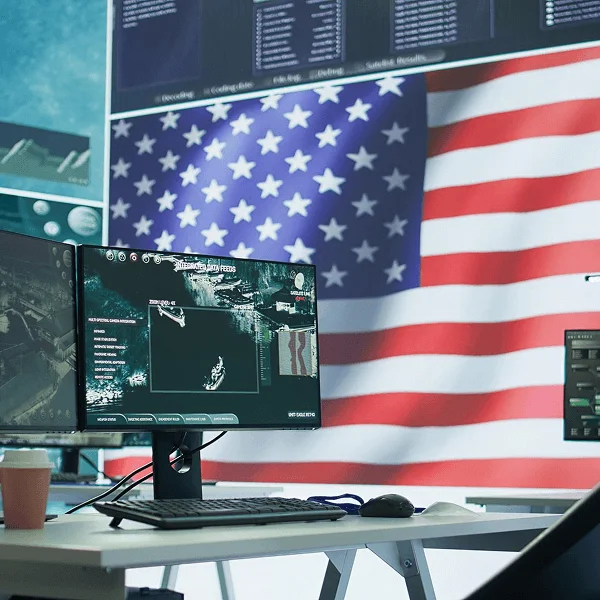 A high-tech workspace featuring dual monitors displaying integrated data feeds, with a prominent U.S. flag in the background.