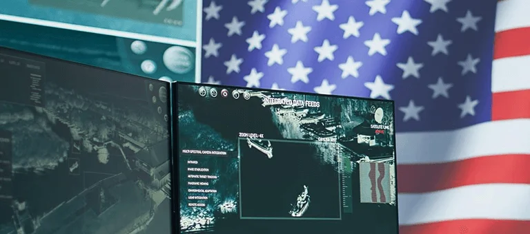 A high-tech workspace featuring dual monitors displaying integrated data feeds, with a prominent U.S. flag in the background.