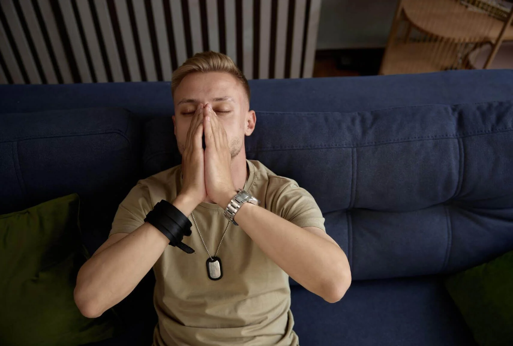 Man sits on couch, hands clasped, wearing tan shirt and dog tags.
