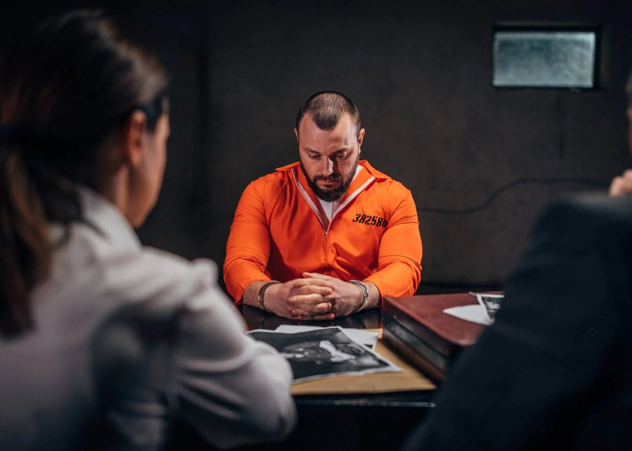 Prisoner in orange jumpsuit sits handcuffed during interrogation with lawyers.
