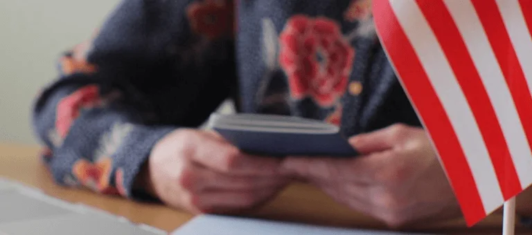 A person in a floral shirt holds a passport, with a small American flag and laptop on the table, symbolizing travel or citizenship.