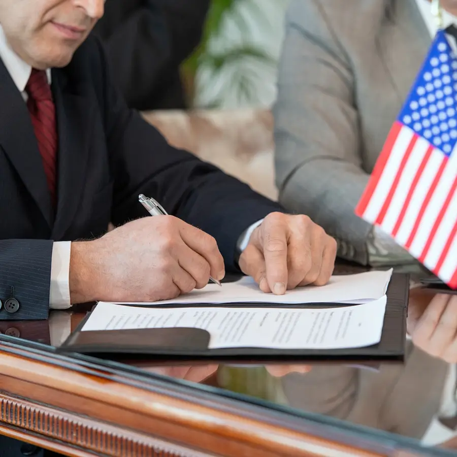 Man signing a document at a table with an American flag, possibly related to a business agreement.