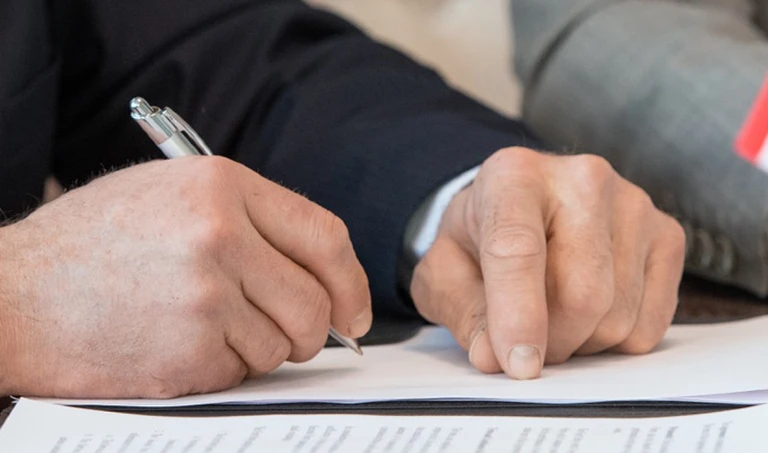 Man signing a document at a table with an American flag, possibly related to a business agreement.