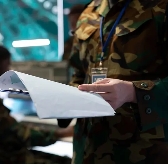Soldier in camouflage uniform holds a clipboard with documents in a command center.